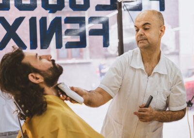 Barber expertly shaving a bearded client in a bustling barbershop.